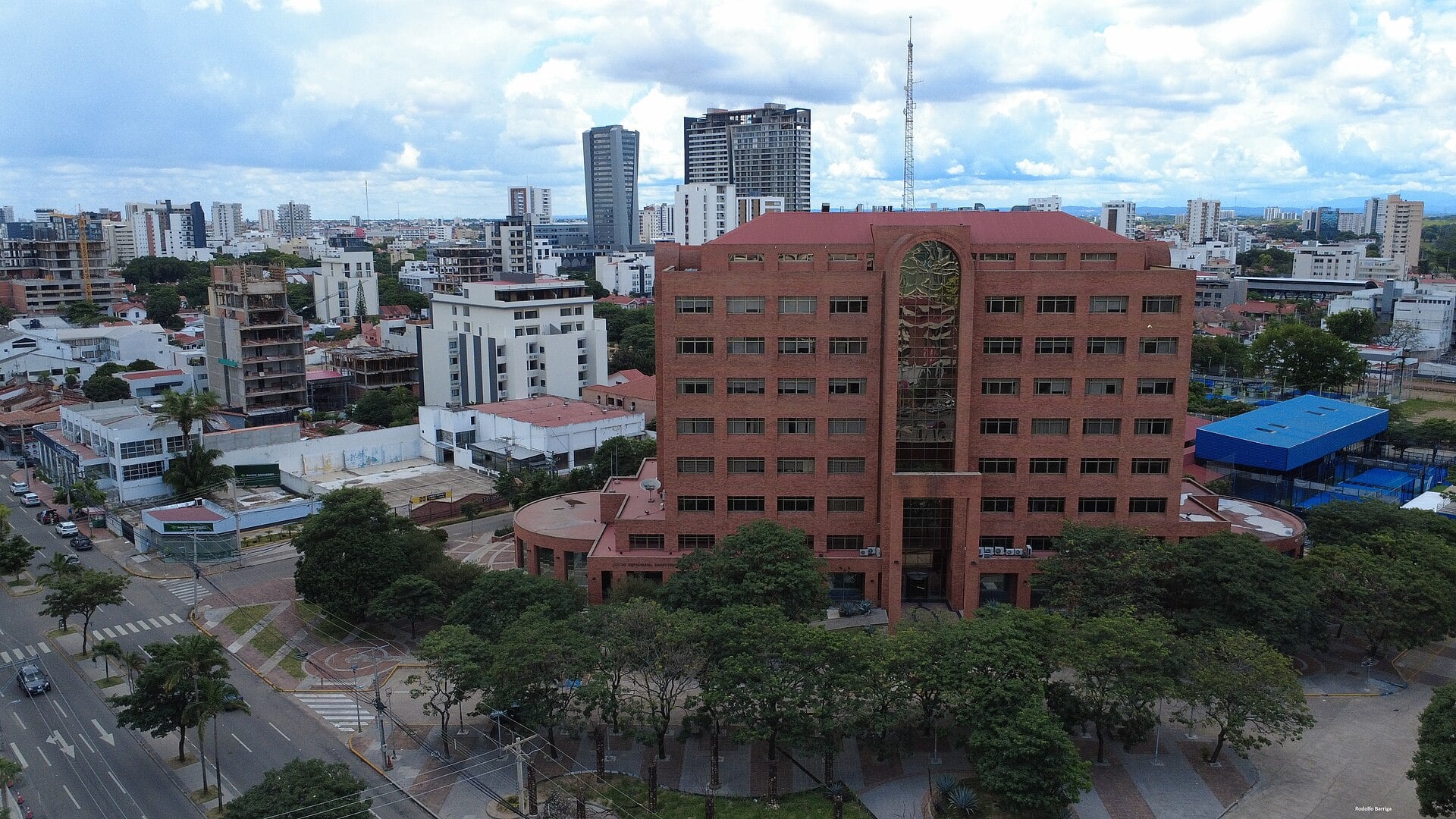 Aerial view of a red-brick office tower surrounded by trees and low buildings, with high-rises and a cloudy sky behind