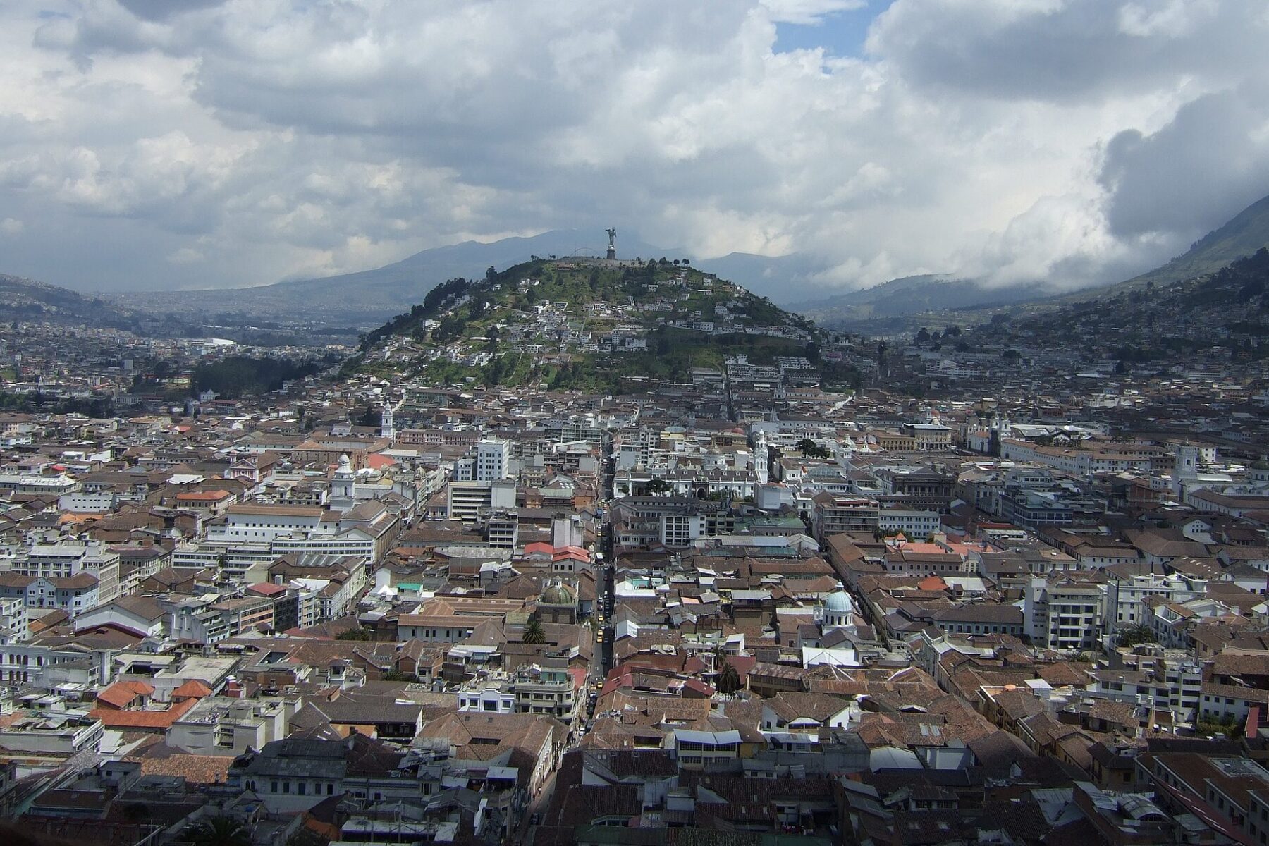 Dense rooftops of central Quito stretching towards a green hill topped by a winged Virgin statue, with Andean mountains and clouds behind
