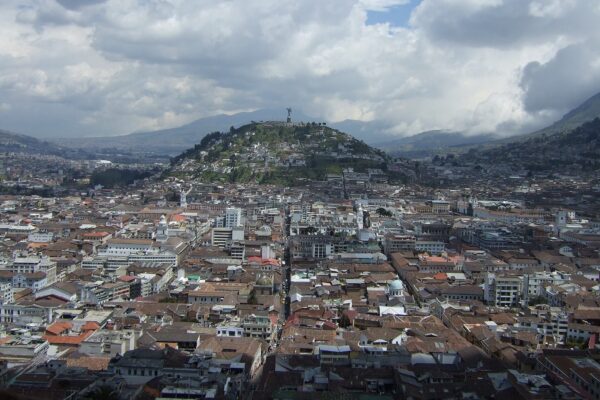 Dense rooftops of central Quito stretching towards a green hill topped by a winged Virgin statue, with Andean mountains and clouds behind