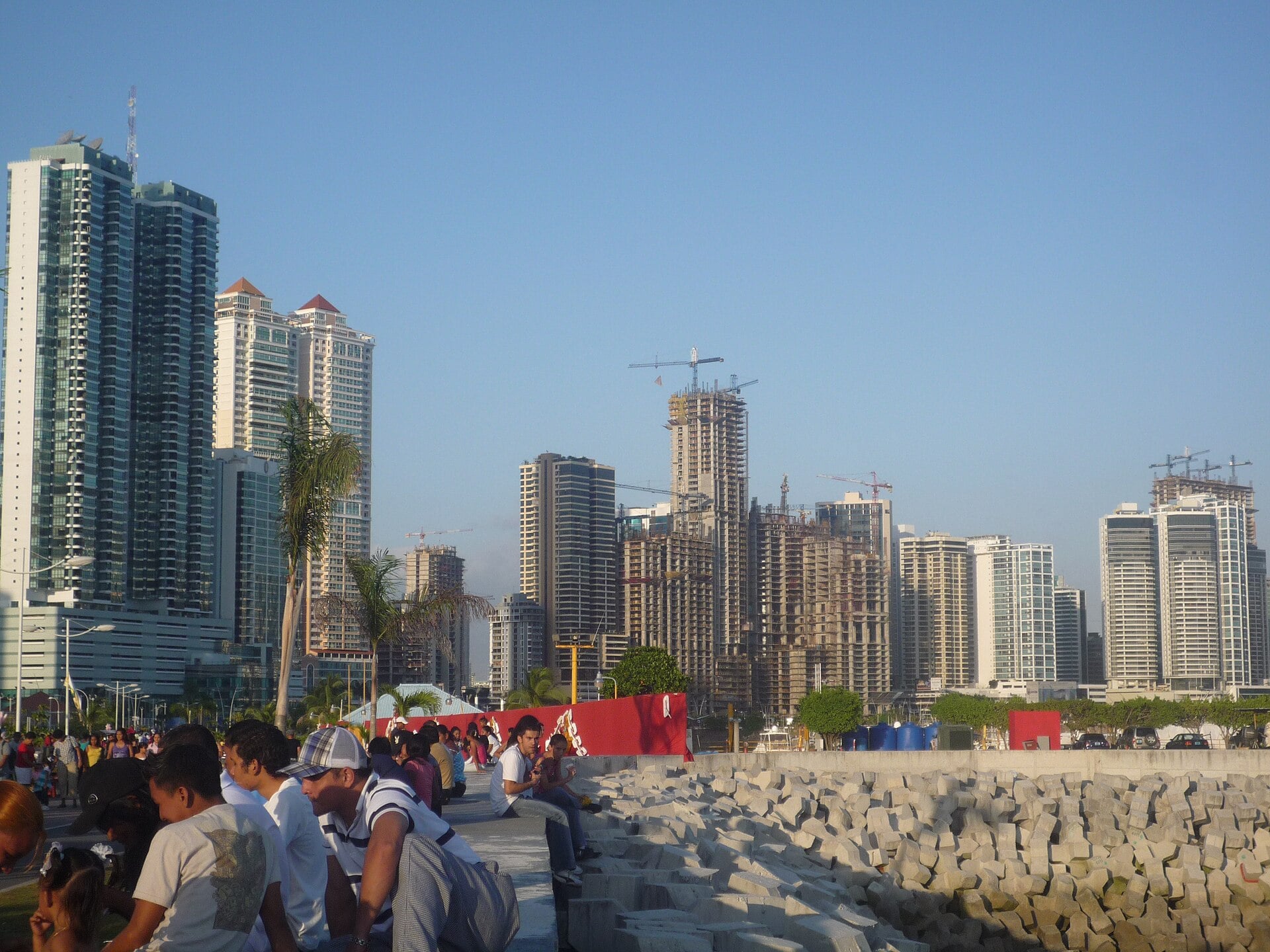 Apartment towers, some still under construction with cranes, on the Panama City skyline under a clear blue sky, with people sitting beside a stone seawall in the foreground