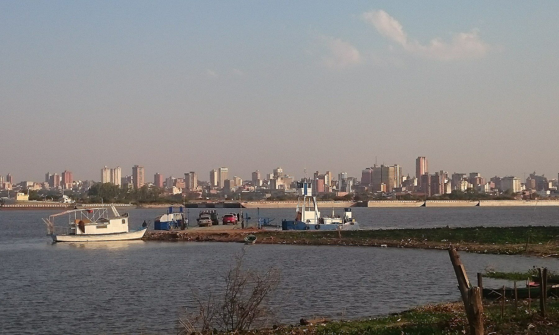 High-rise buildings of Asunción on the far bank of a wide river, with a small white boat, a ferry and cars at a riverside landing in the foreground