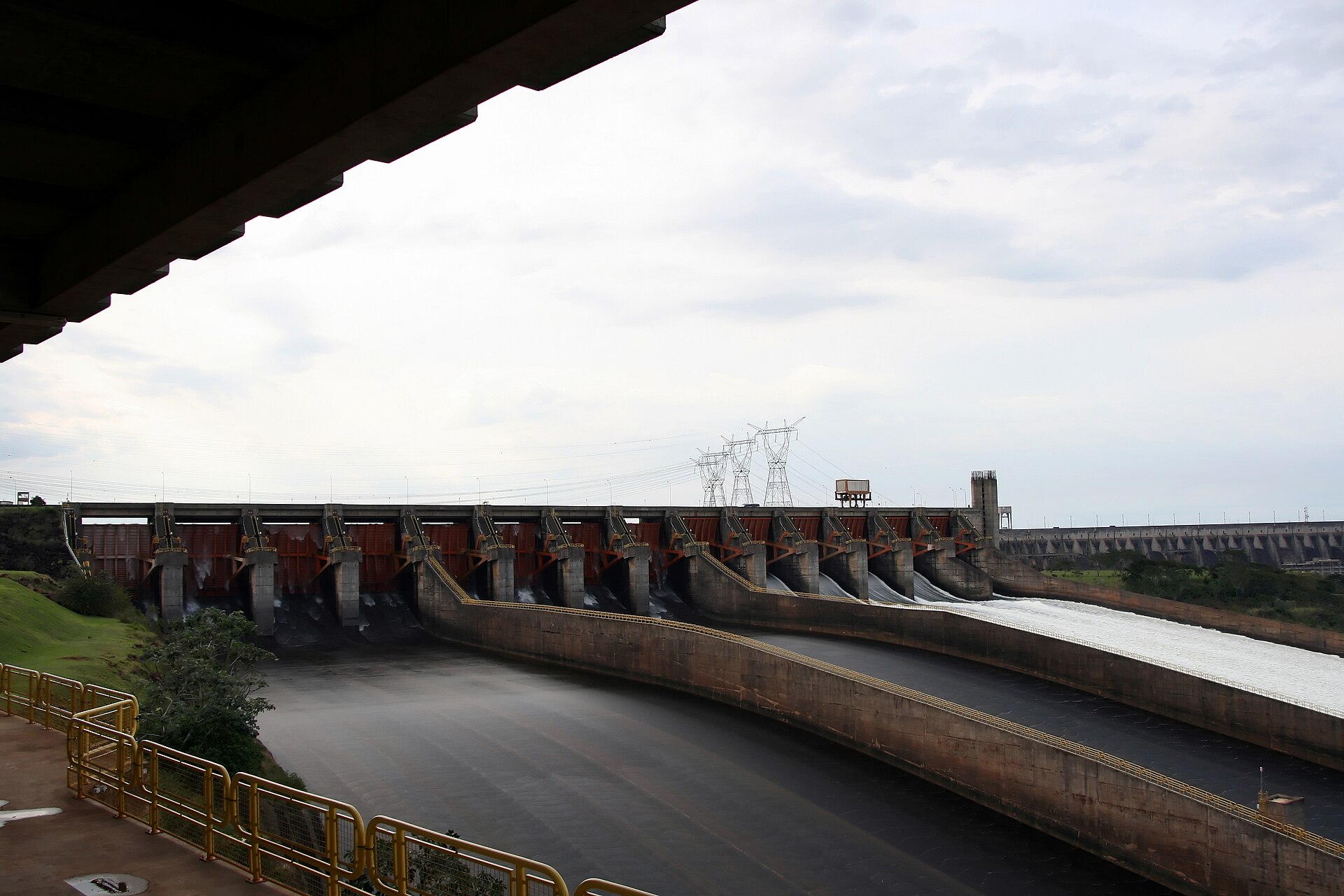 The concrete spillway of the Itaipu dam with its row of gates and three curved chutes, water rushing down the right-hand chute, under an overcast sky