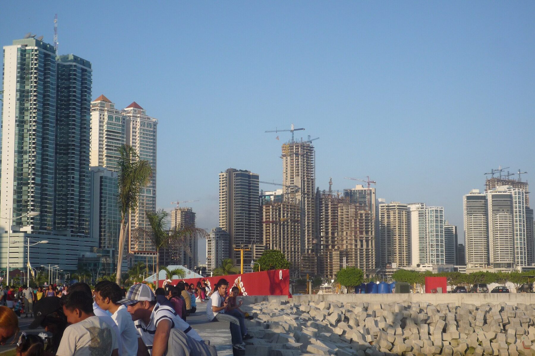 Apartment towers, some still under construction with cranes, on the Panama City skyline under a clear blue sky, with people sitting beside a stone seawall in the foreground