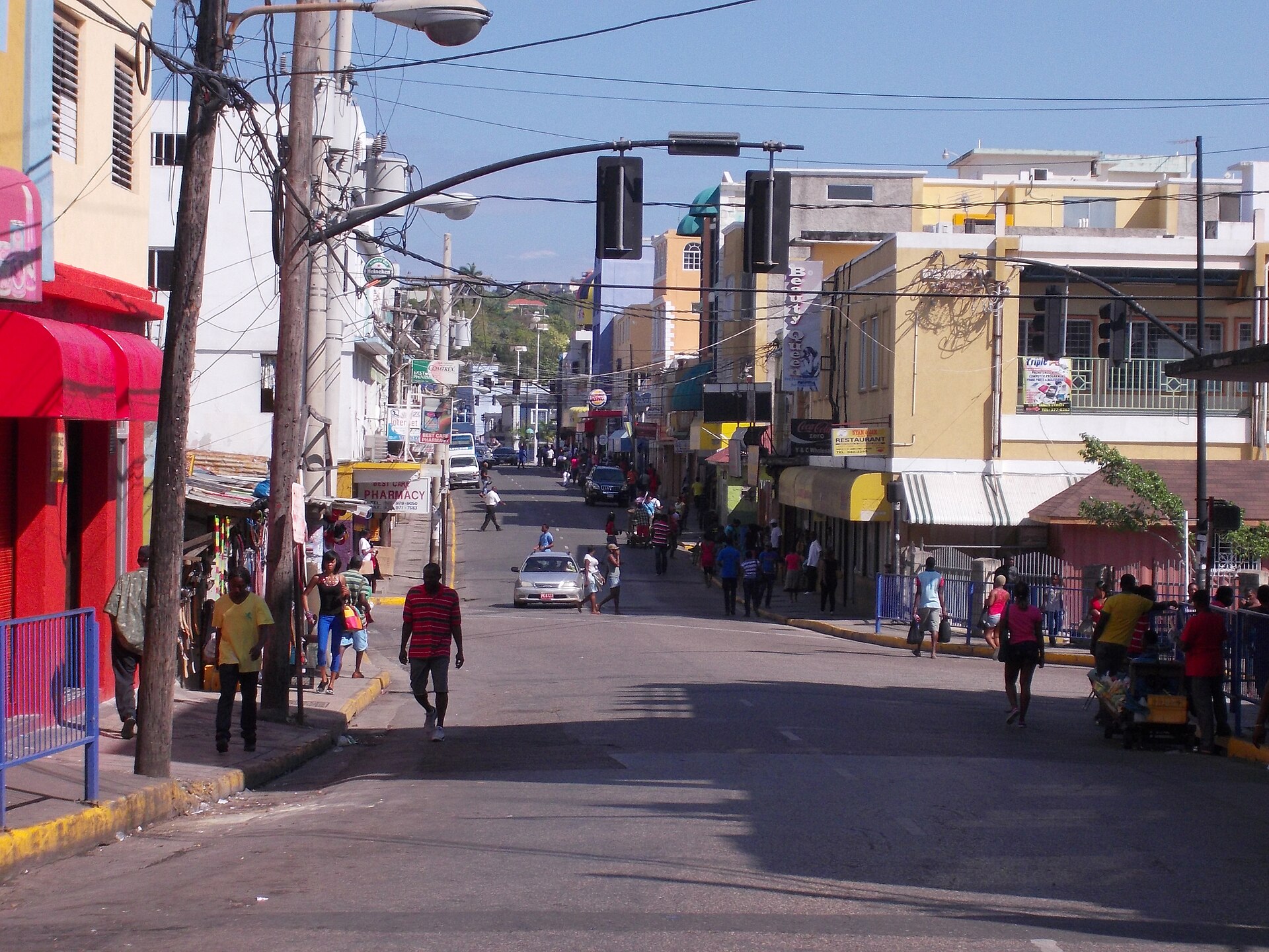 A sunny street in Montego Bay with pedestrians, shops with red and yellow awnings, traffic lights and overhead wires