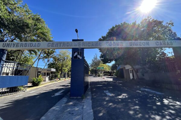 A blue gate arch reading Universidad Centroamericana José Simeón Cañas spans a road lined with palm and shade trees under a bright sun