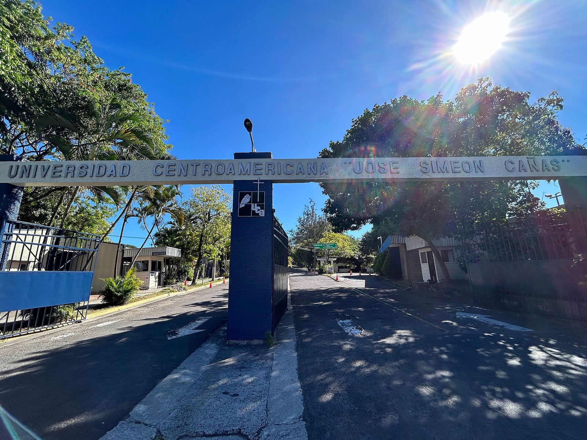 A blue gate arch reading Universidad Centroamericana José Simeón Cañas spans a road lined with palm and shade trees under a bright sun