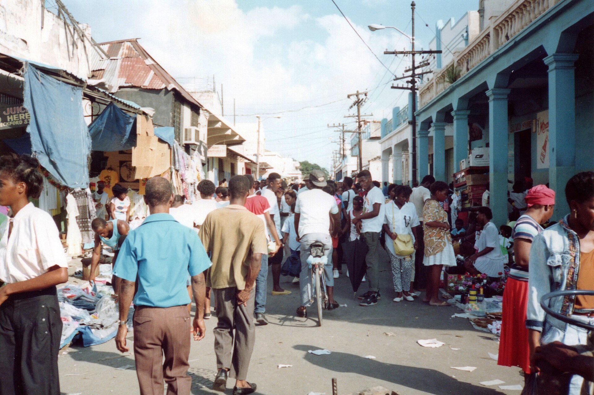 A crowded market street lined with stalls and colonnaded shopfronts, with shoppers walking and a man pushing a bicycle