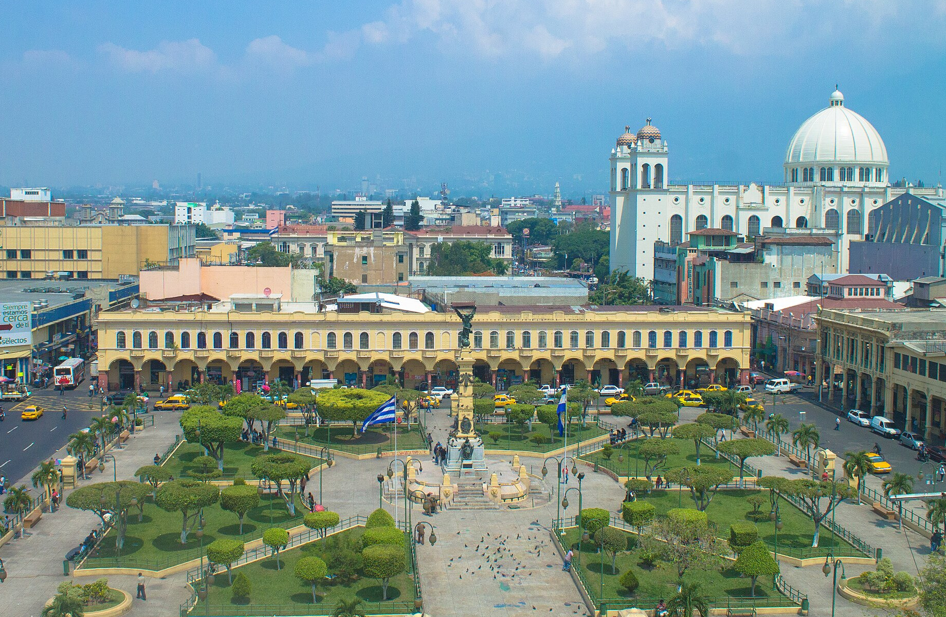 Elevated view of a tree-lined square with a central column monument and a blue-and-white flag, arcaded yellow buildings behind and a white domed church on the right