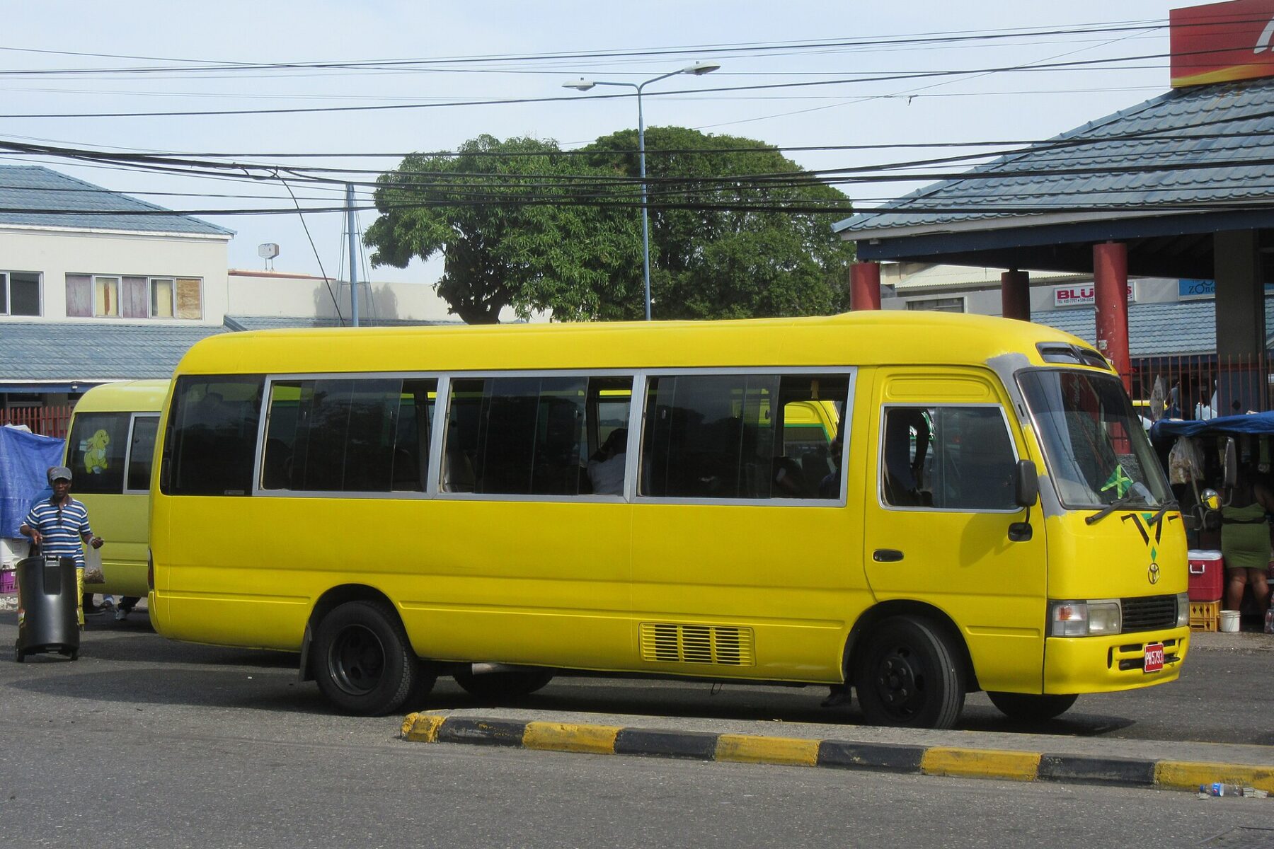 A yellow Toyota Coaster minibus stopped at a kerb beside a covered terminal in Kingston, with a second yellow bus, a street lamp and trees behind it