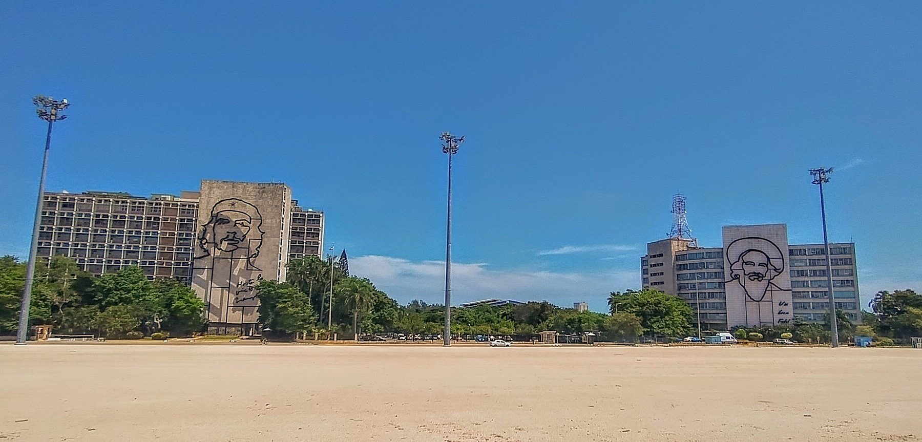 A wide sandy plaza under a blue sky with two government buildings among trees, each carrying a large steel outline portrait