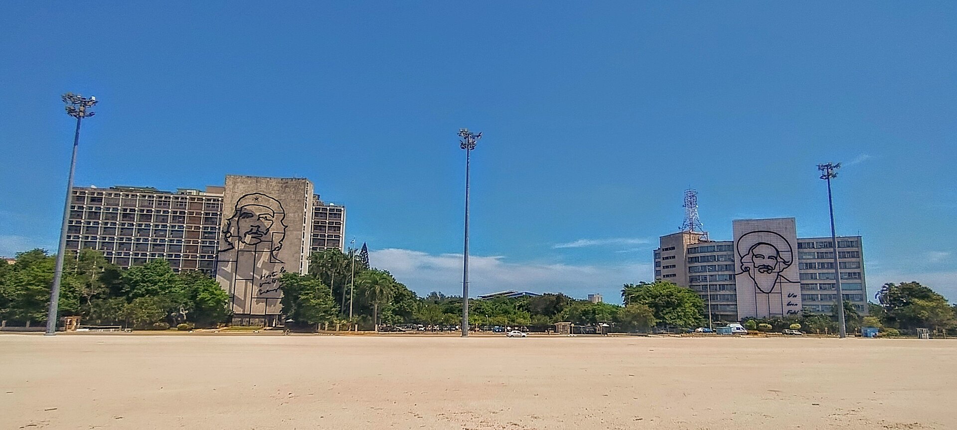 A wide sandy plaza under a blue sky with two government buildings among trees, each carrying a large steel outline portrait