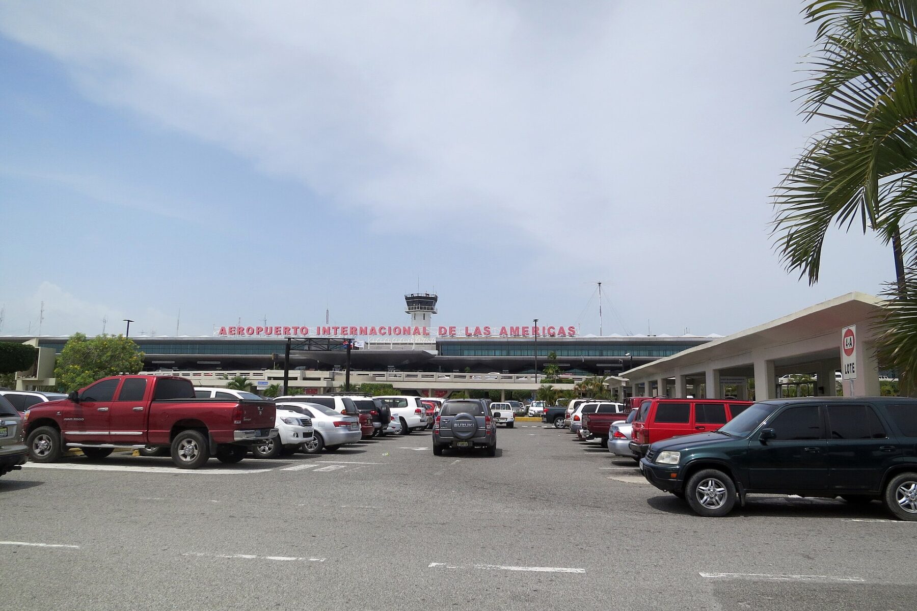 Front of Las Américas International Airport in Santo Domingo with the red terminal sign, control tower, parked cars and a palm frond
