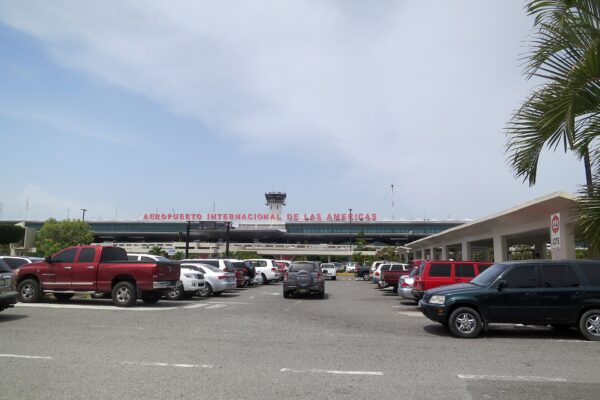Front of Las Américas International Airport in Santo Domingo with the red terminal sign, control tower, parked cars and a palm frond