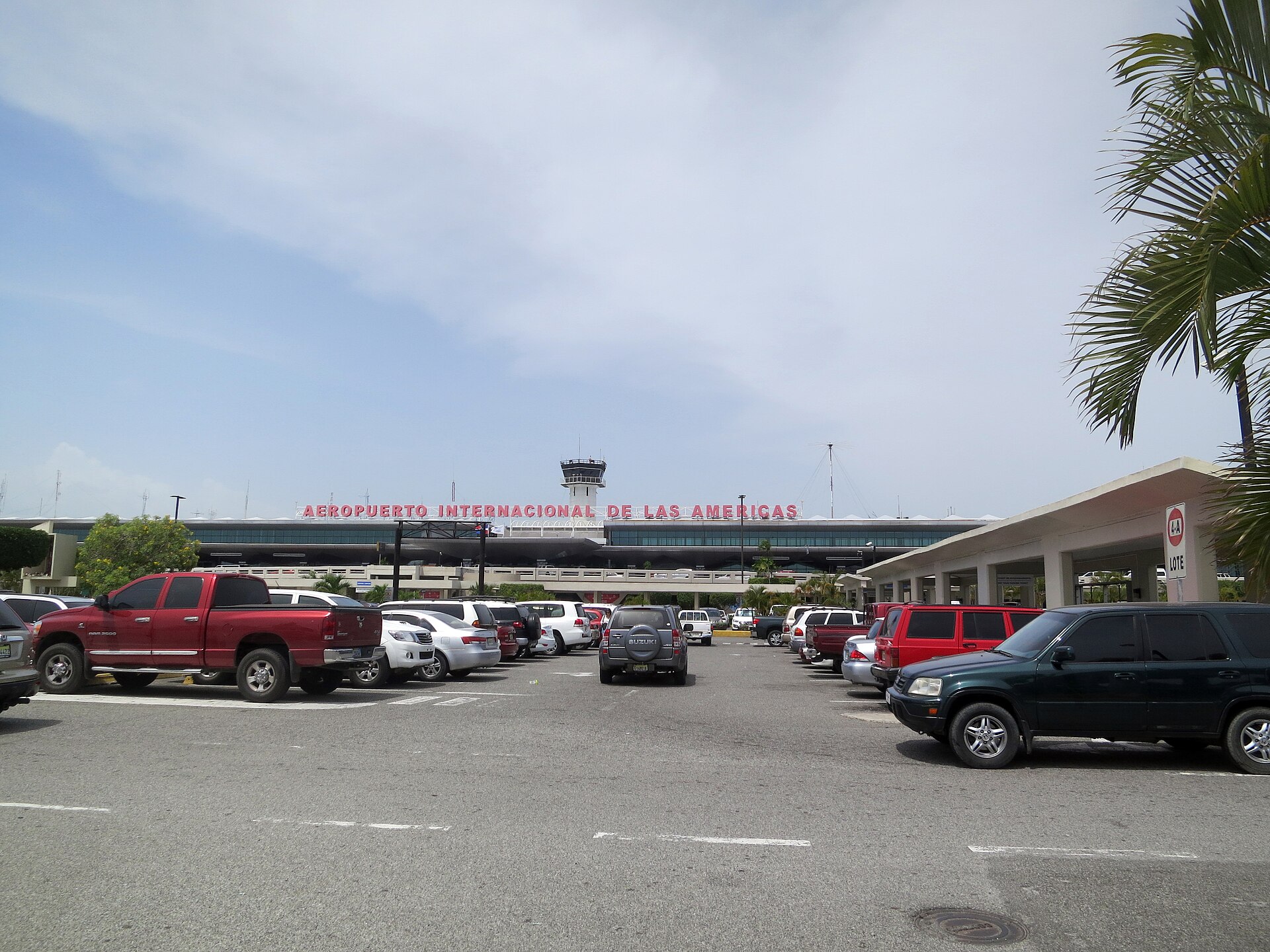 Front of Las Américas International Airport in Santo Domingo with the red terminal sign, control tower, parked cars and a palm frond