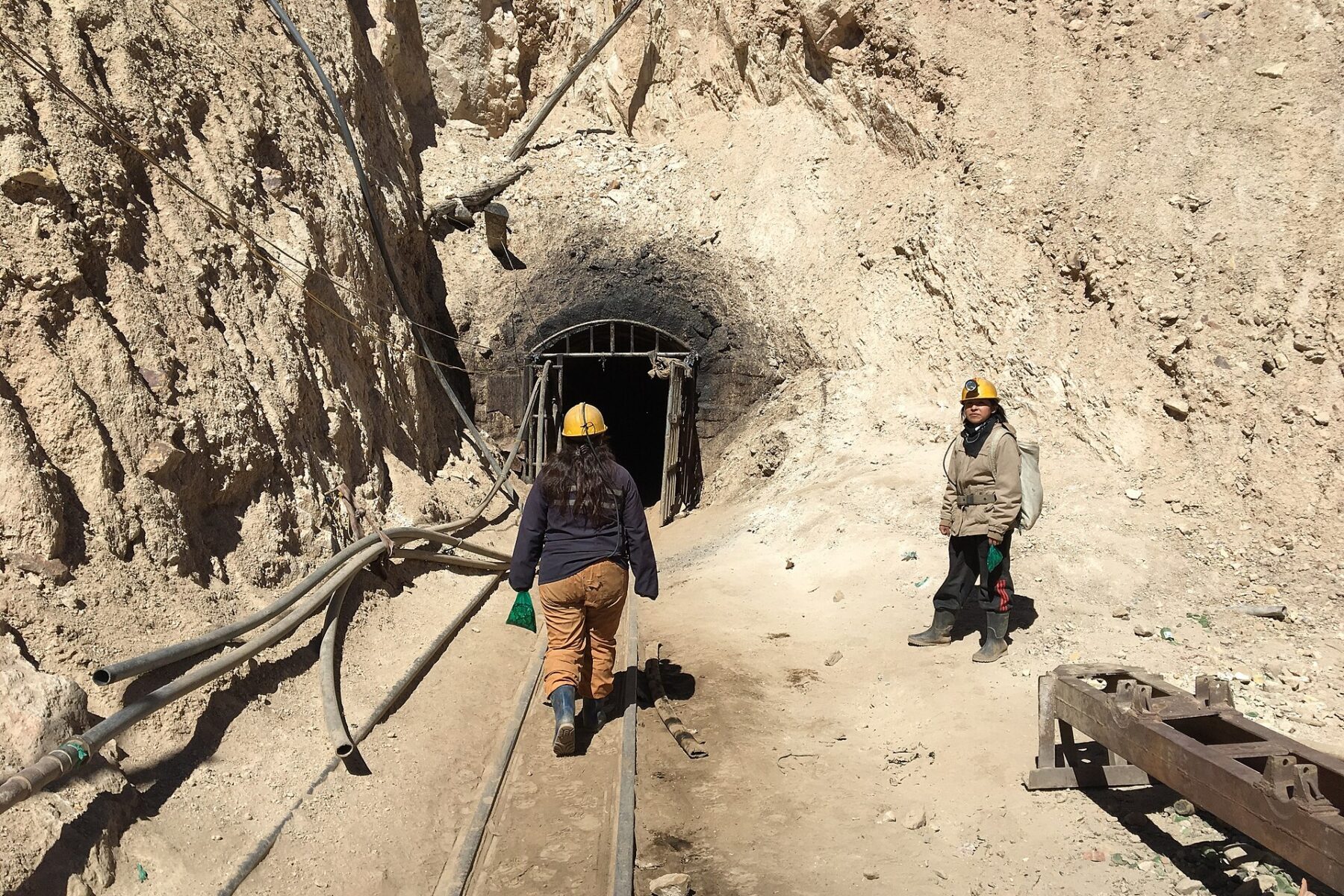Two people in yellow helmets on rail tracks leading to a tunnel entrance cut into a dusty hillside
