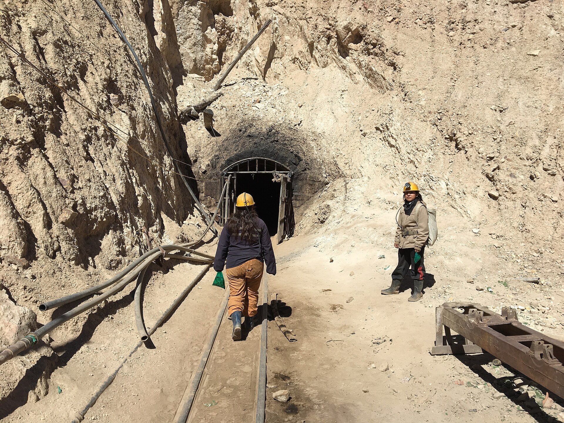 Two people in yellow helmets on rail tracks leading to a tunnel entrance cut into a dusty hillside