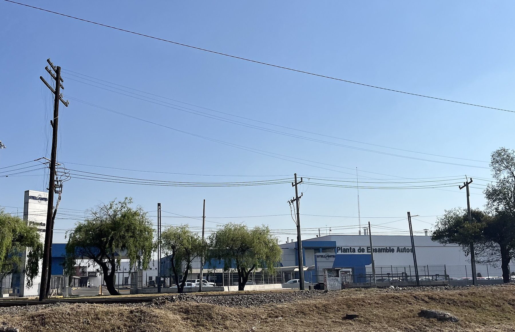 Exterior of a car assembly plant in Toluca, Mexico, behind power lines and willow trees under a clear sky