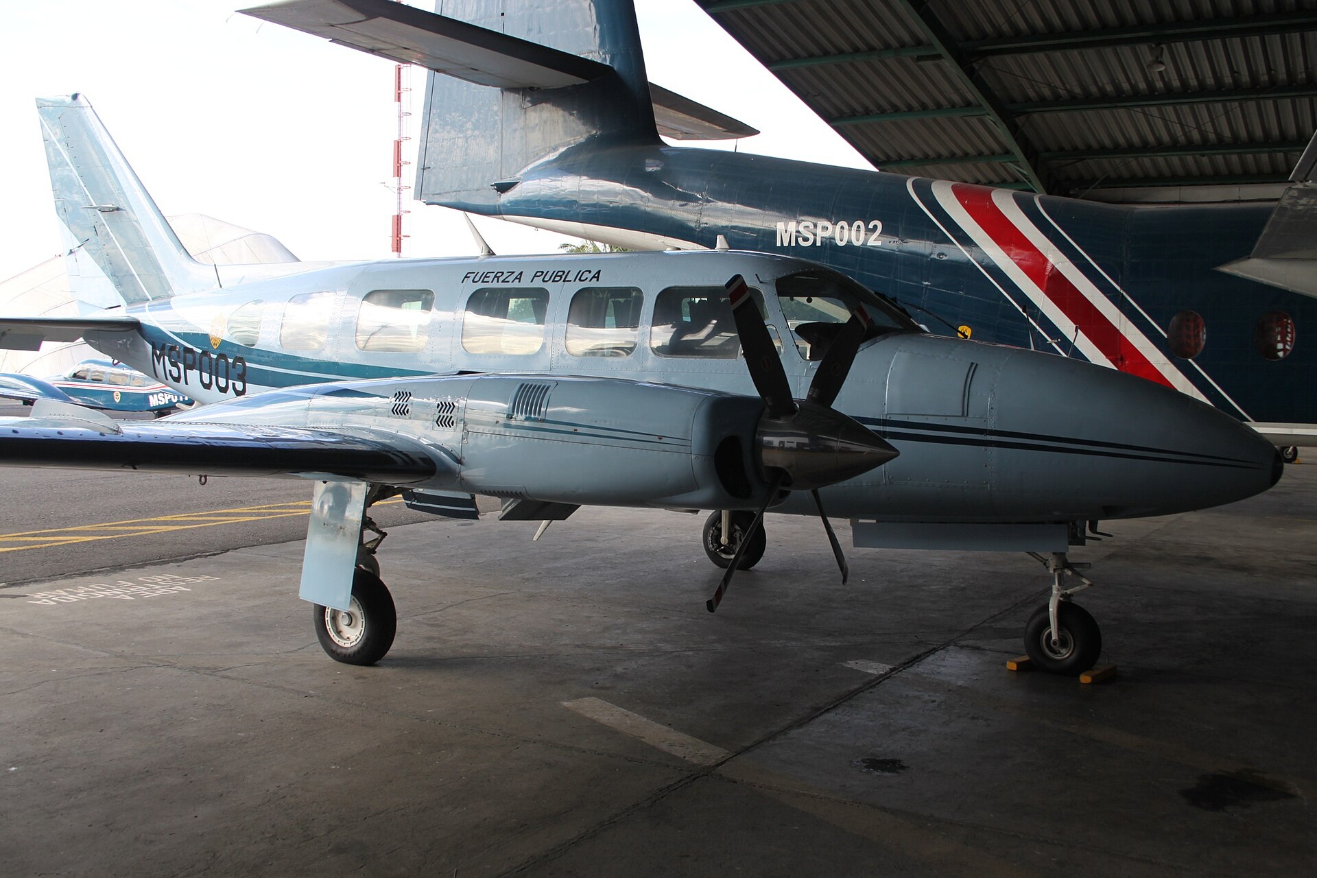 A pale blue twin-engine propeller aircraft marked Fuerza Publica and MSP003 parked in a hangar beside a larger blue aircraft marked MSP002