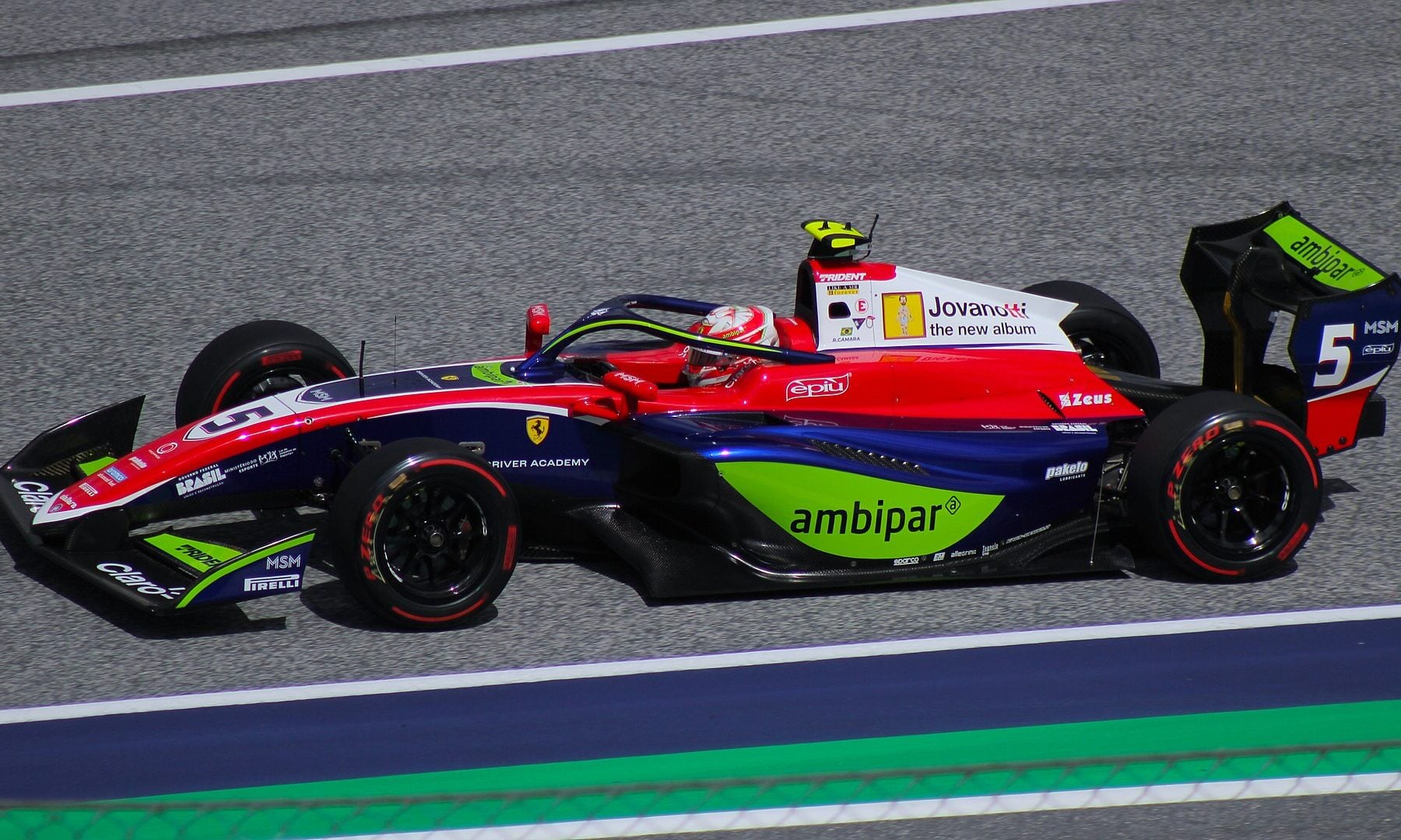 A blue, red and white Trident Formula 3 car with the number 5, a Ferrari Driver Academy logo and green sponsor panels drives along a grey track as the driver in a red and white helmet looks ahead
