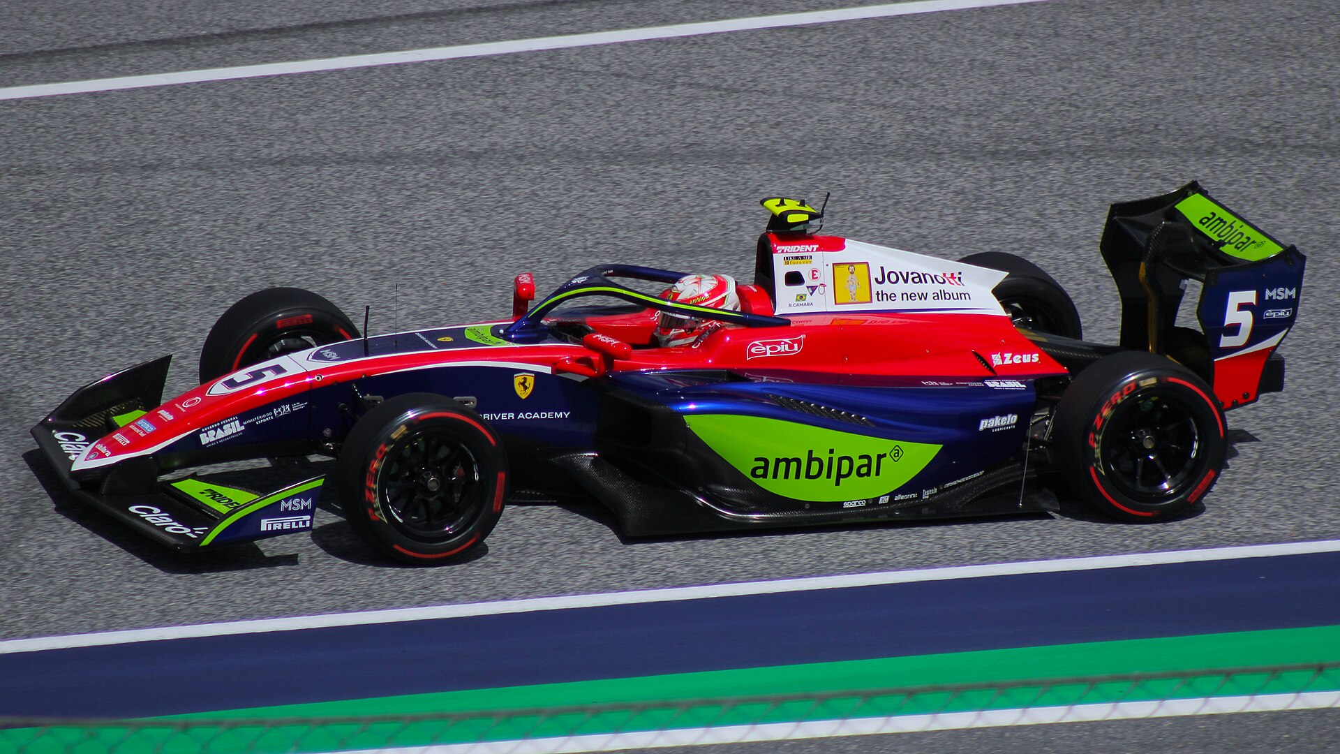 A blue, red and white Trident Formula 3 car with the number 5, a Ferrari Driver Academy logo and green sponsor panels drives along a grey track as the driver in a red and white helmet looks ahead
