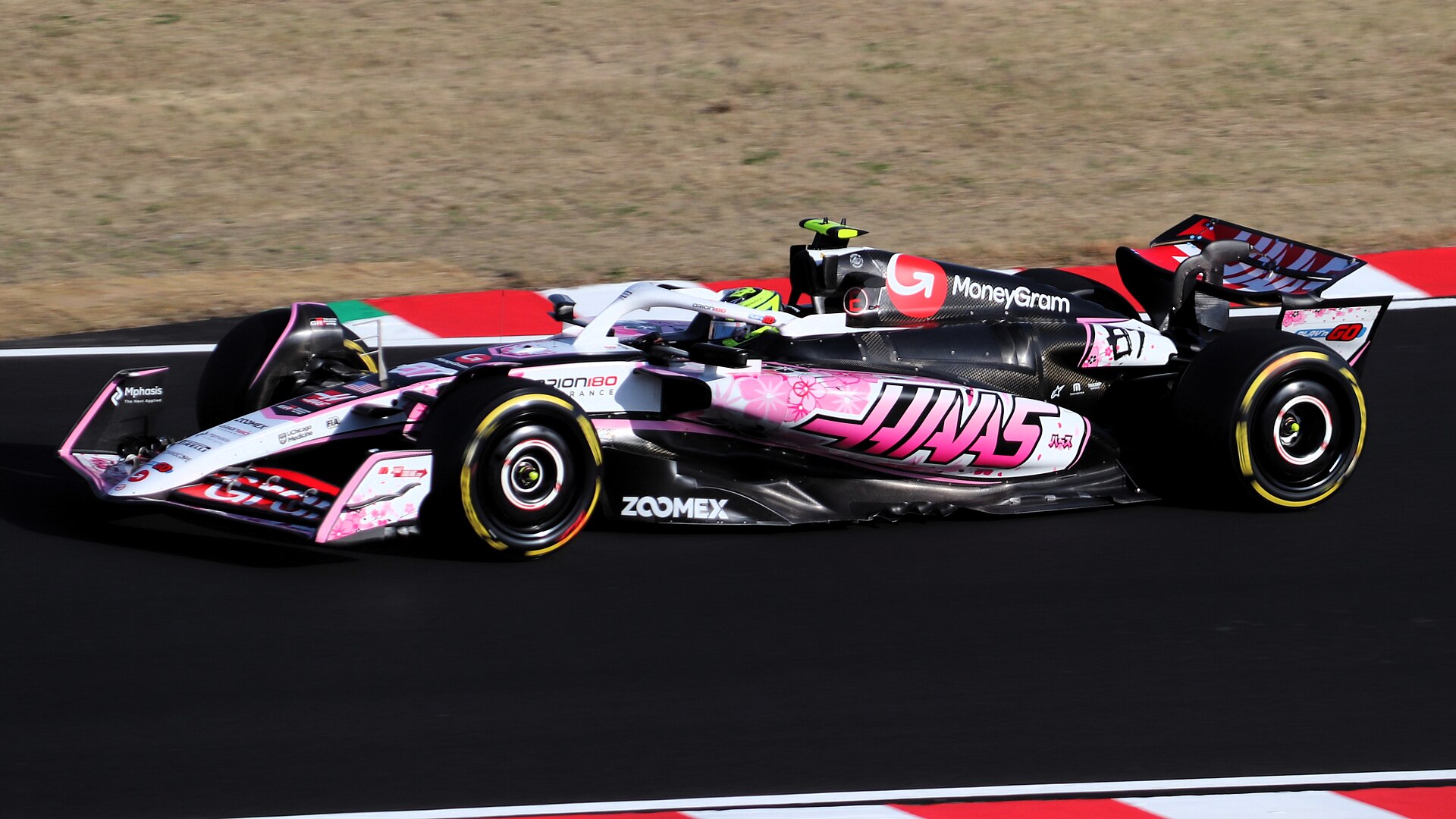 A Haas Formula 1 car in a pink and white special livery with the number 87 drives along a black track past red and white kerbs