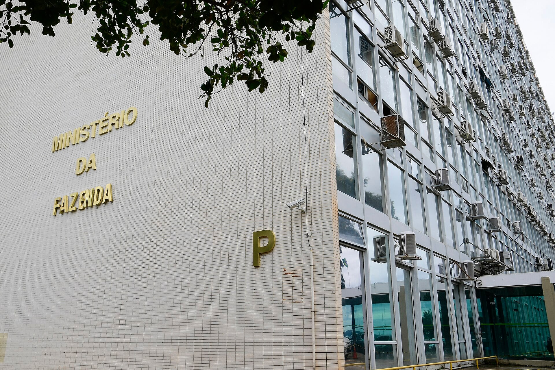 A white tiled wall and glass facade of a government building with gold letters reading Ministério da Fazenda and a large letter P, with tree branches at top left