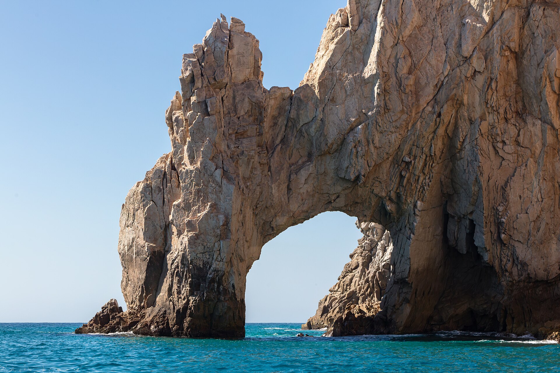 A tall, weathered rock arch rising from turquoise sea water under a clear blue sky