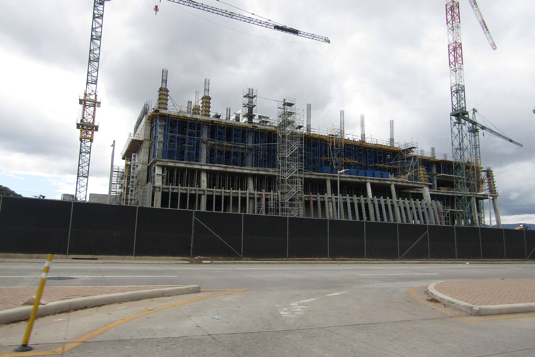 A multi-storey concrete building under construction, with scaffolding and two tower cranes, behind a black fence beside an empty road