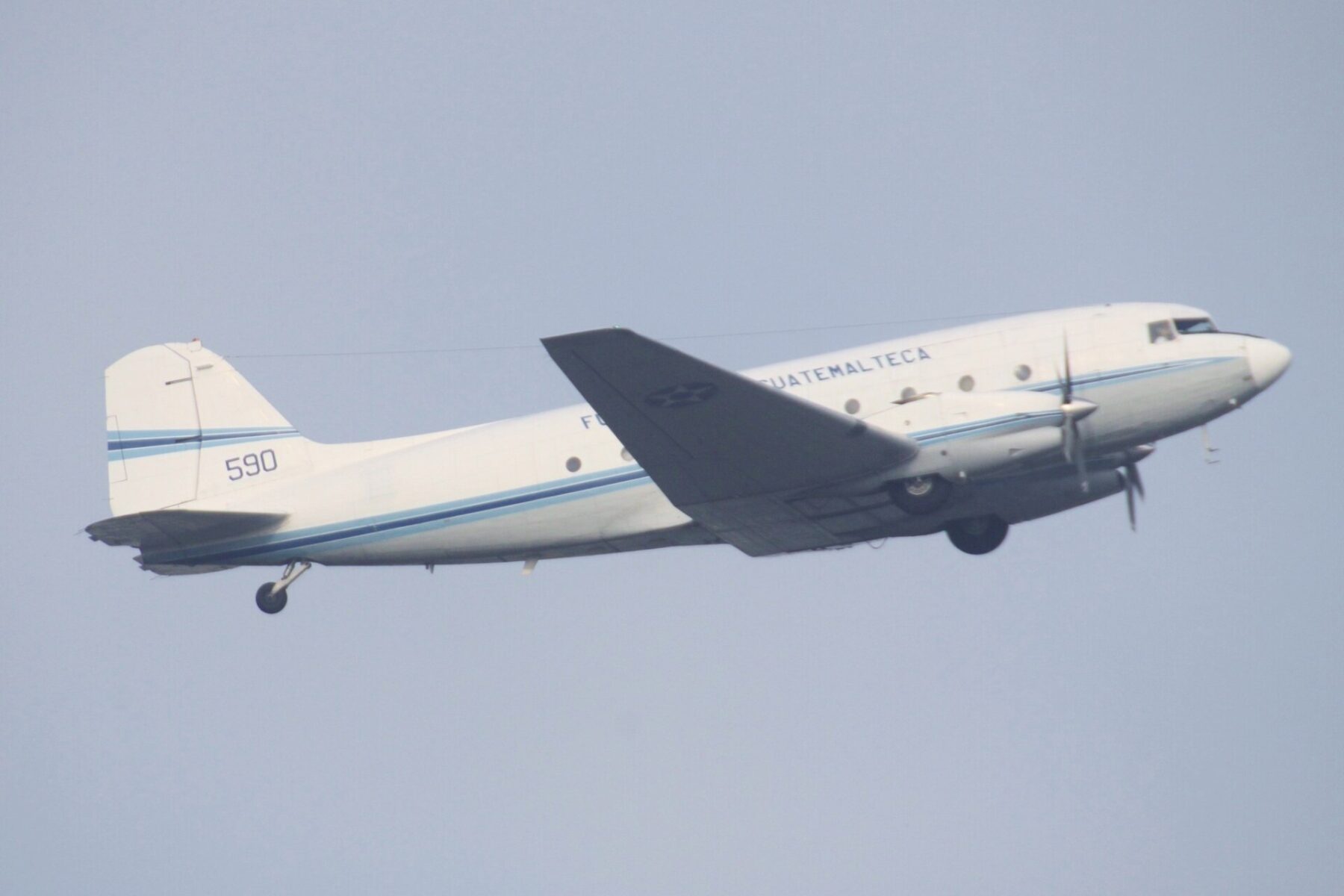 A white Guatemalan Air Force Douglas DC-3 transport, number 590, climbing after take-off against a pale sky