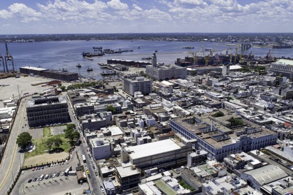 Aerial view of the old city of Montevideo with low rooftops, port cranes at the left, ships and the grey-blue River Plate stretching to the horizon under a cloudy sky