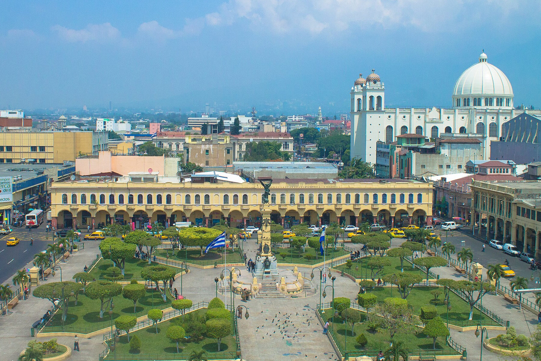 Elevated view of a tree-lined square with a central column monument and a blue-and-white flag, arcaded yellow buildings behind and a white domed church on the right
