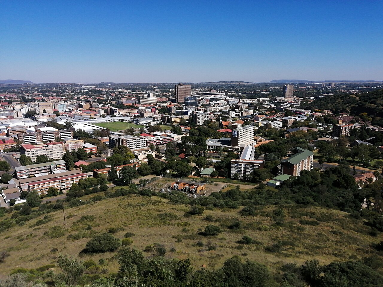 Central Bloemfontein, South Africa, seen from a hillside, with office towers, low-rise buildings and trees under a blue sky