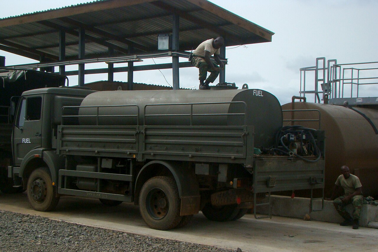A green Armed Forces of Liberia fuel truck at a motor pool with one soldier standing on the tank and another seated nearby