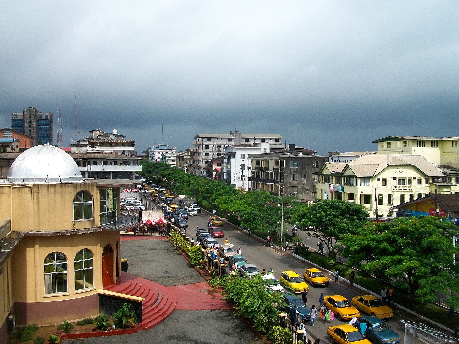 A tree-lined street in central Monrovia with yellow taxis, traffic and low buildings under a dark rain sky