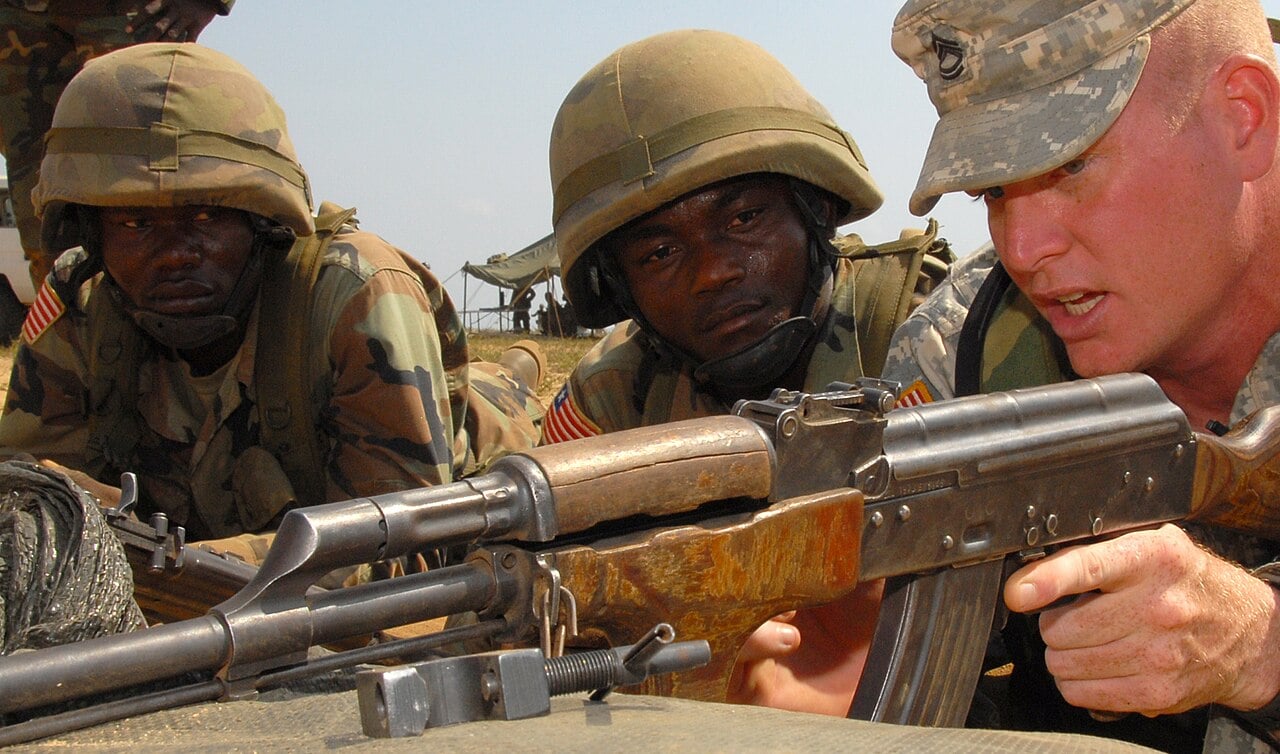 A US Army mentor shows two Liberian soldiers in helmets how to aim an AK-47 rifle at a firing range