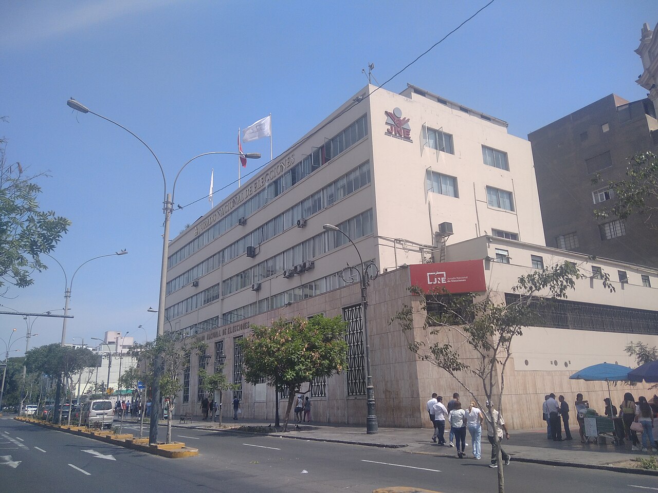 The white headquarters of Peru's National Elections Jury in central Lima, with the JNE logo and flags on the roof and pedestrians on the pavement