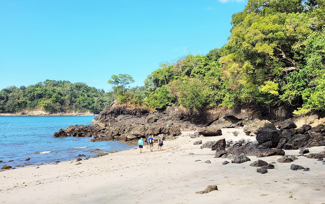 Visitors walk along a sandy cove below forested rocks in Manuel Antonio National Park, Costa Rica.