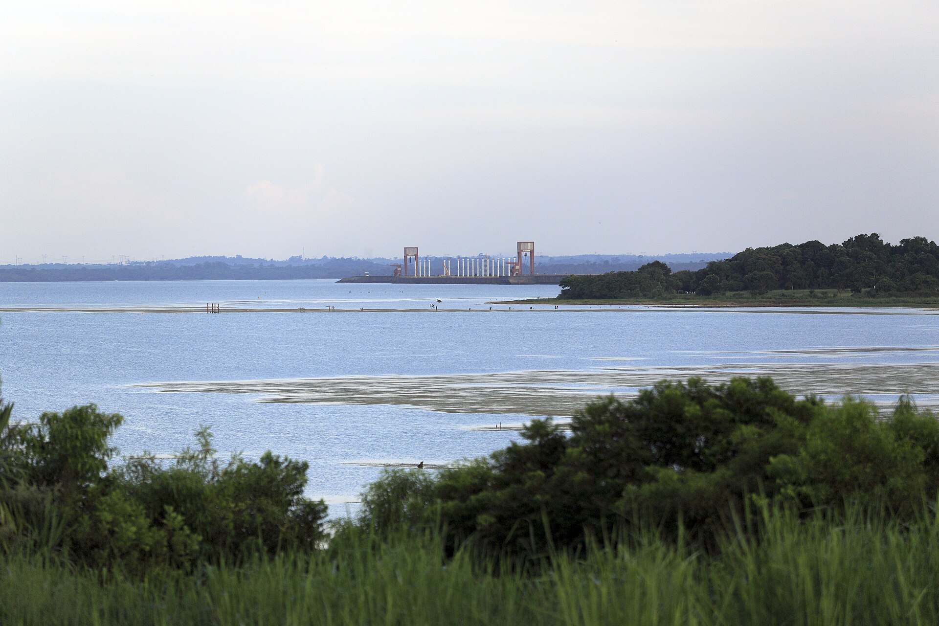 A wide, pale reservoir under a hazy sky with the Itaipu dam and its portal cranes visible as a small structure on the far shore, grass in the foreground
