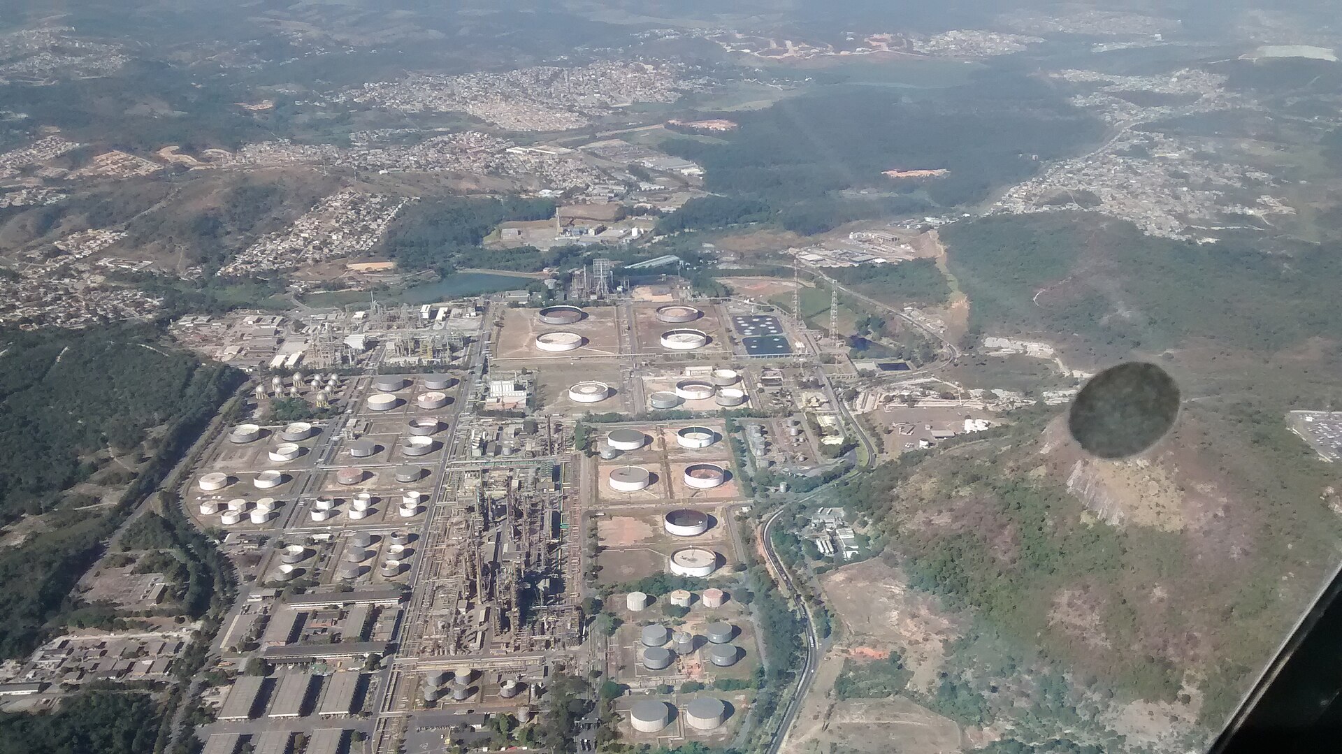 Aerial view of a large oil refinery with round storage tanks and process units among green hills and suburbs