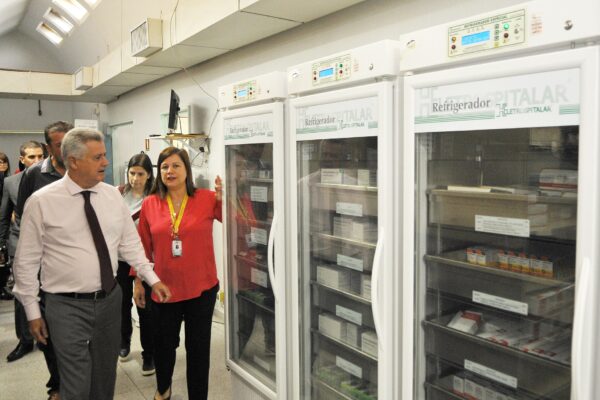 Two officials and staff walk past three tall hospital refrigerators holding boxes of medicines in a hospital pharmacy in Brasilia