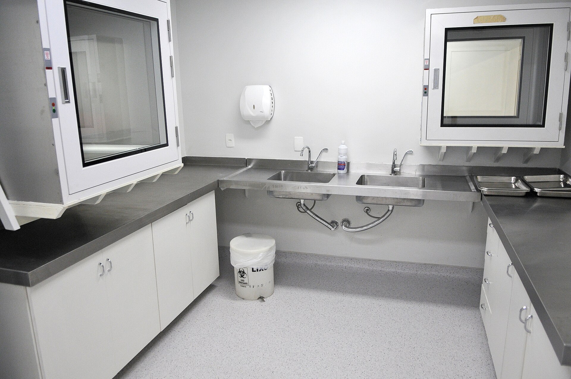 A clean hospital preparation room with stainless steel counters, two sinks, white cabinets, two pass-through hatches and a biohazard waste bin