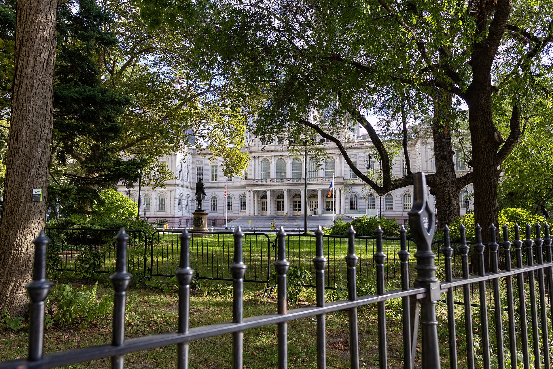 New York City Hall, a pale stone building with a flagpole, seen through trees and an iron fence in City Hall Park