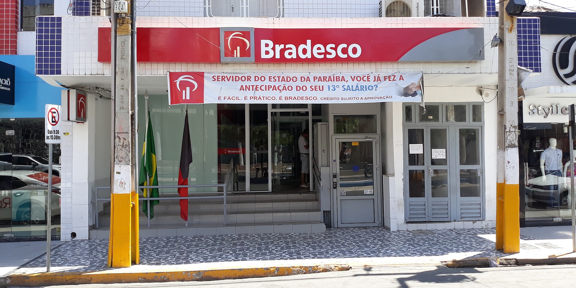 Front of a red-and-white Bradesco bank branch in Sousa, Paraíba, with a banner about 13th-salary advances and Brazilian and state flags at the door
