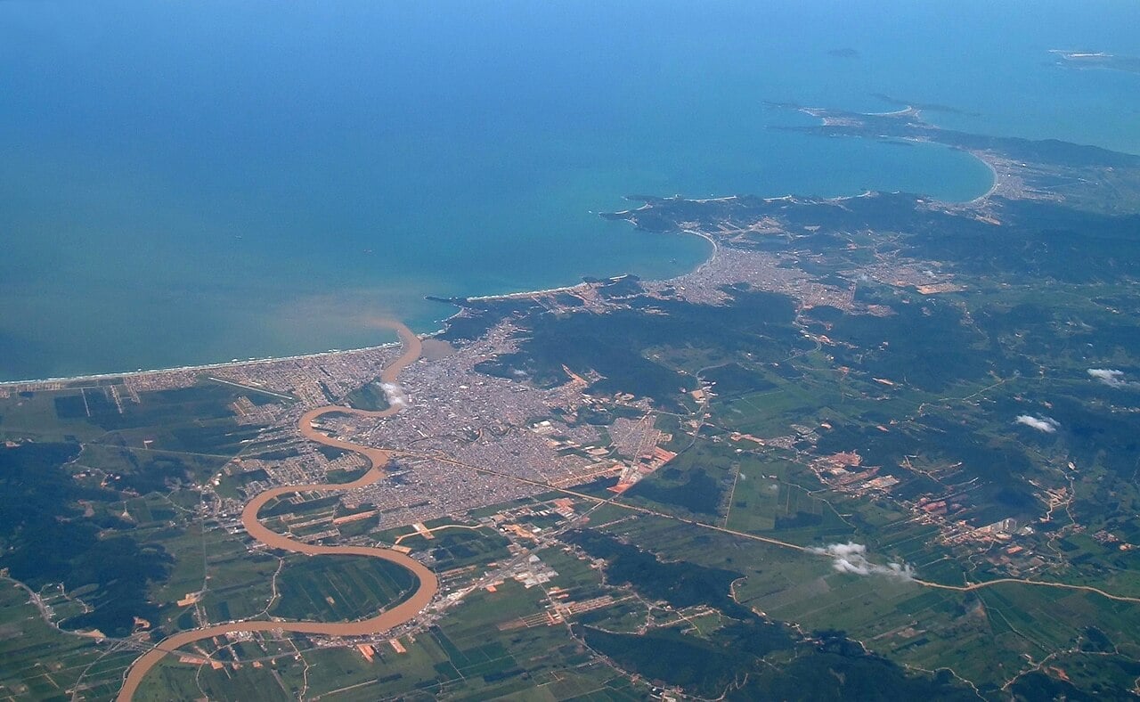Aerial view of the city of Itajaí at the mouth of a winding brown river meeting a blue sea, with green hills and farmland inland