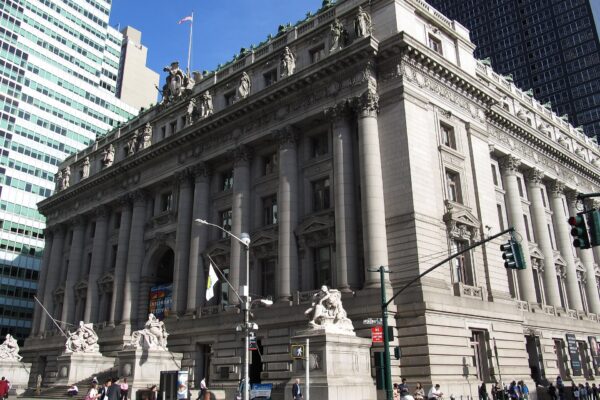 The granite facade of the Alexander Hamilton U.S. Custom House in Lower Manhattan with sculpted figures along the roofline under a blue sky