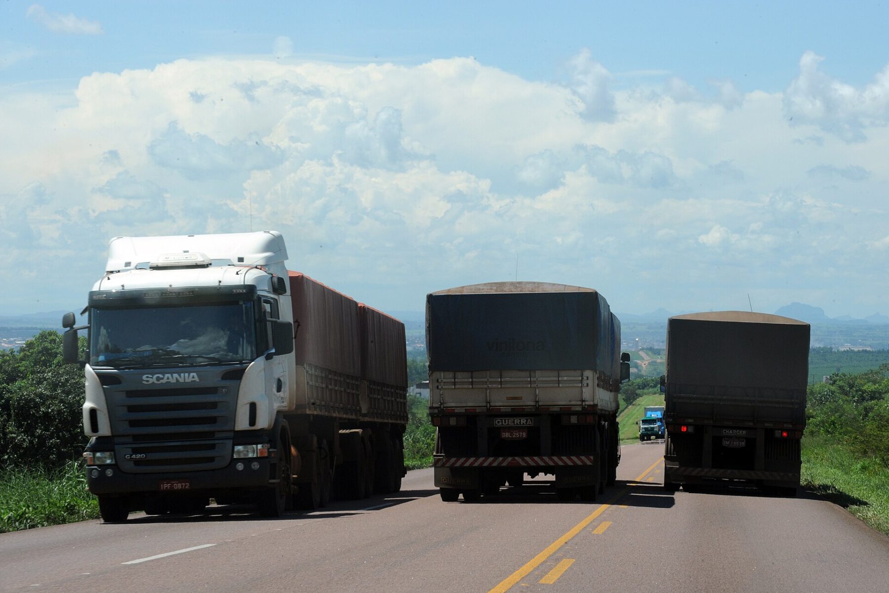 Three loaded grain trucks on a highway with green hills and a cloudy blue sky in the background near Rondonópolis, Mato Grosso
