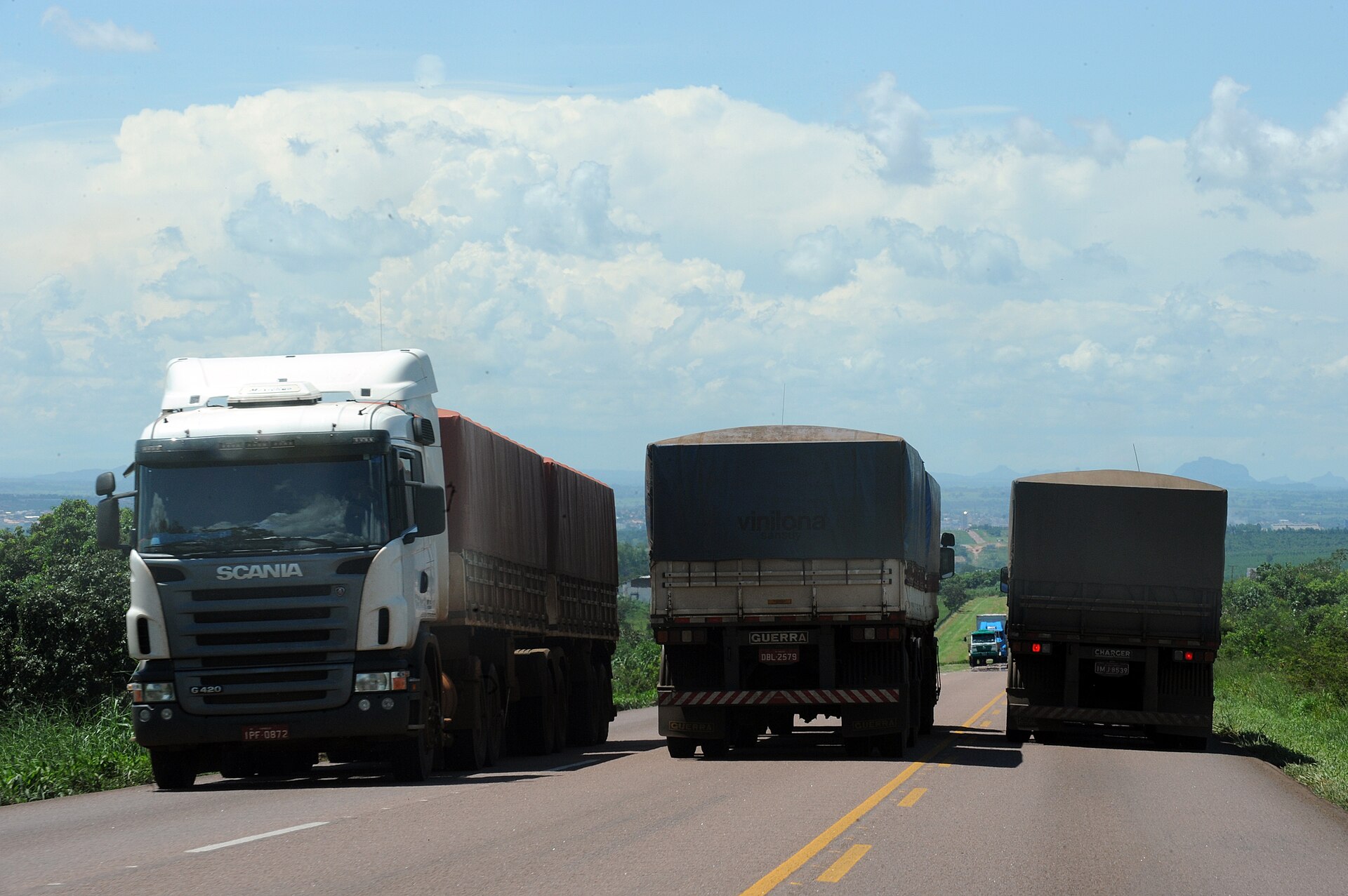 Three loaded grain trucks on a highway with green hills and a cloudy blue sky in the background near Rondonópolis, Mato Grosso