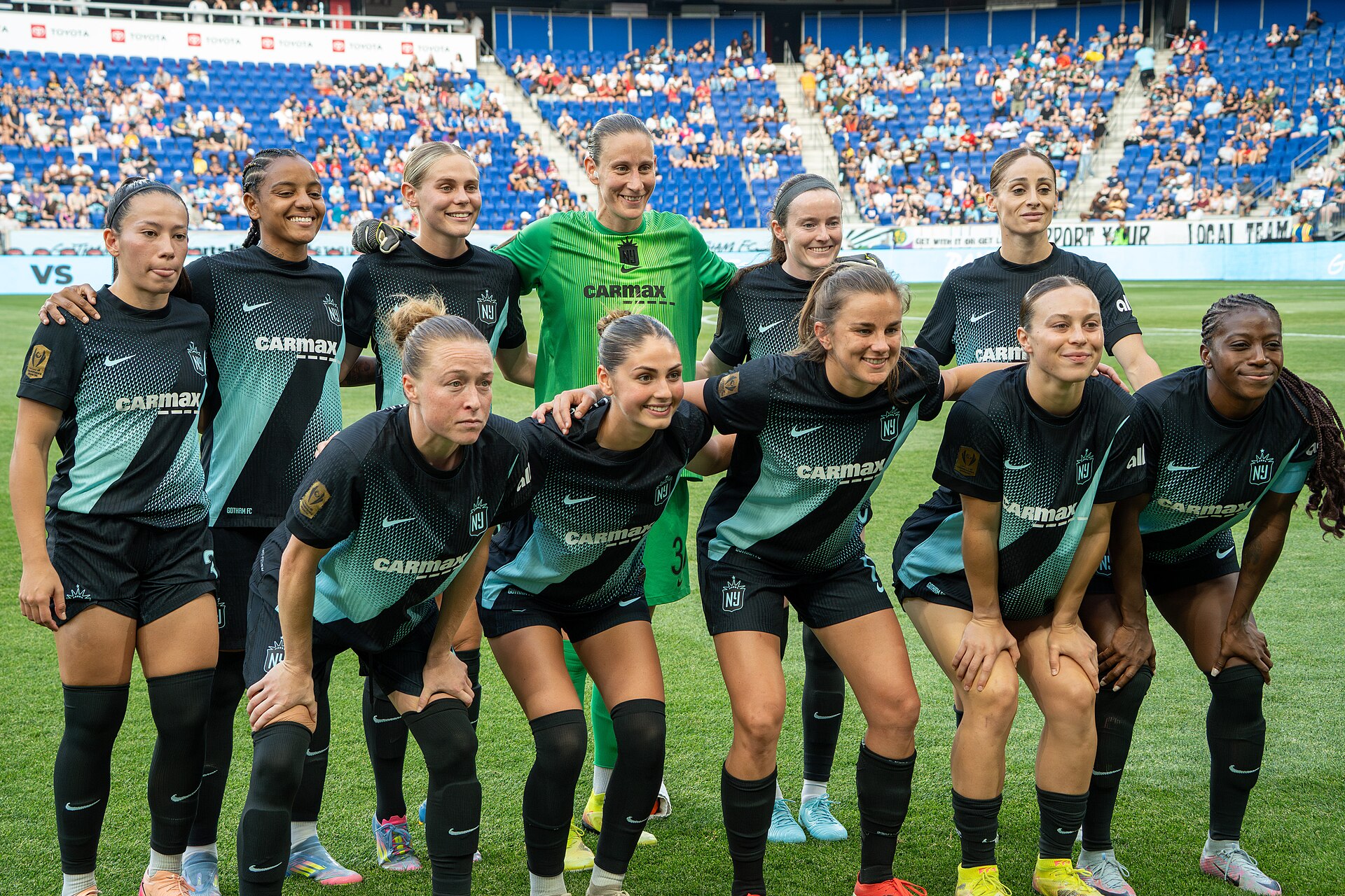 Gotham FC players in dark and teal kits and a goalkeeper in green posing together on the pitch before a match, with fans in blue seats behind