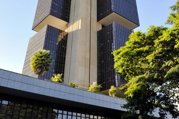 The dark glass towers and stone core of the Central Bank of Brazil headquarters in Brasilia seen from below, with trees in the foreground
