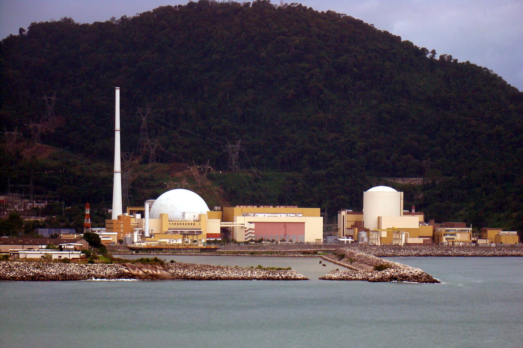 The Angra nuclear power complex at Angra dos Reis, with a white containment dome and a tall chimney in front of a forested hill beside the sea